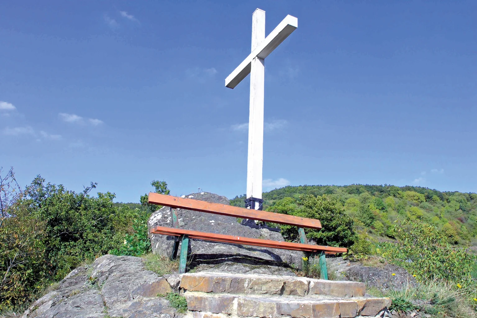 Weißes Kreuz auf einem Felsen in Altenahr, darunter steht eine rote Bank.