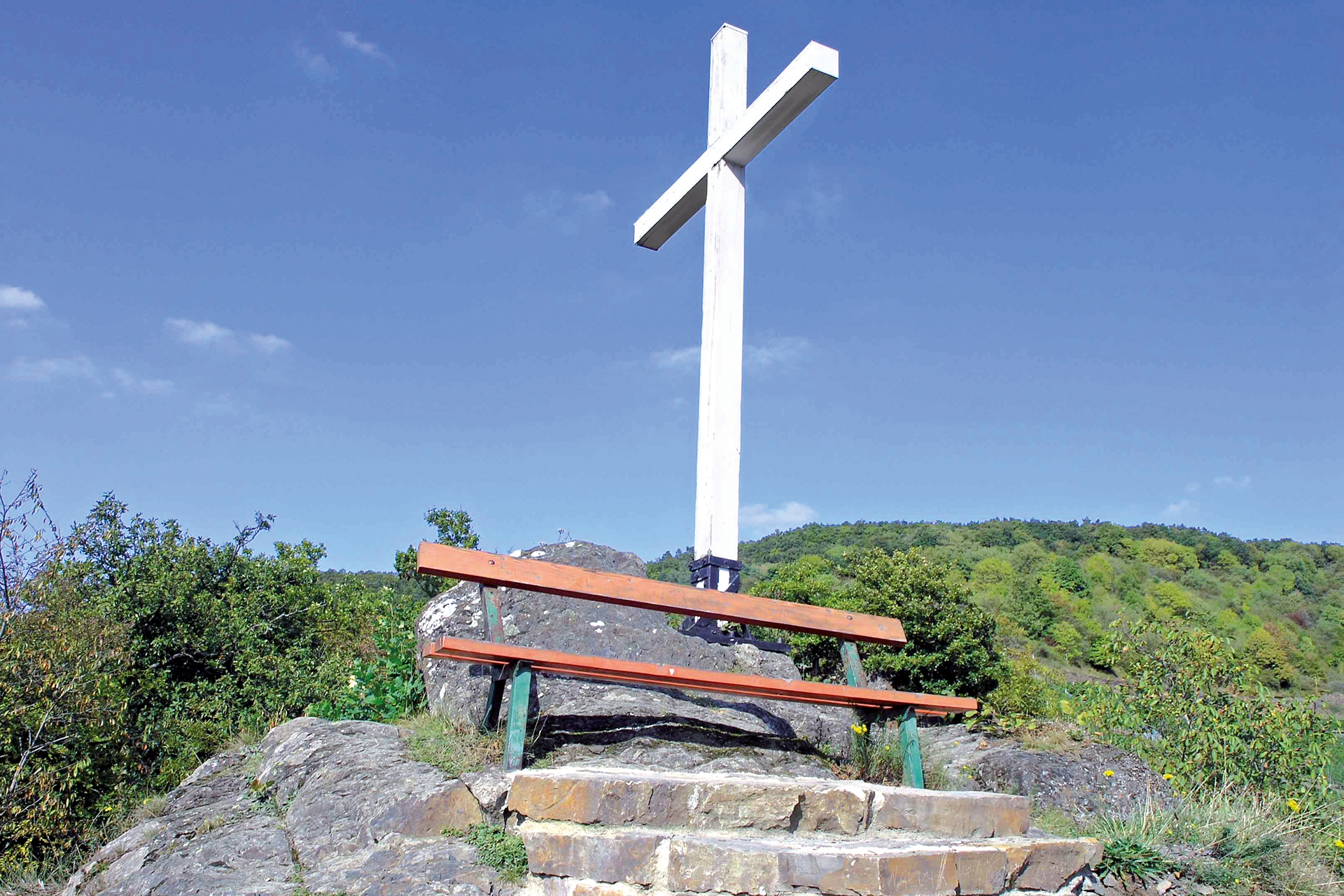 Weißes Kreuz auf einem Felsen in Altenahr, darunter steht eine rote Bank.